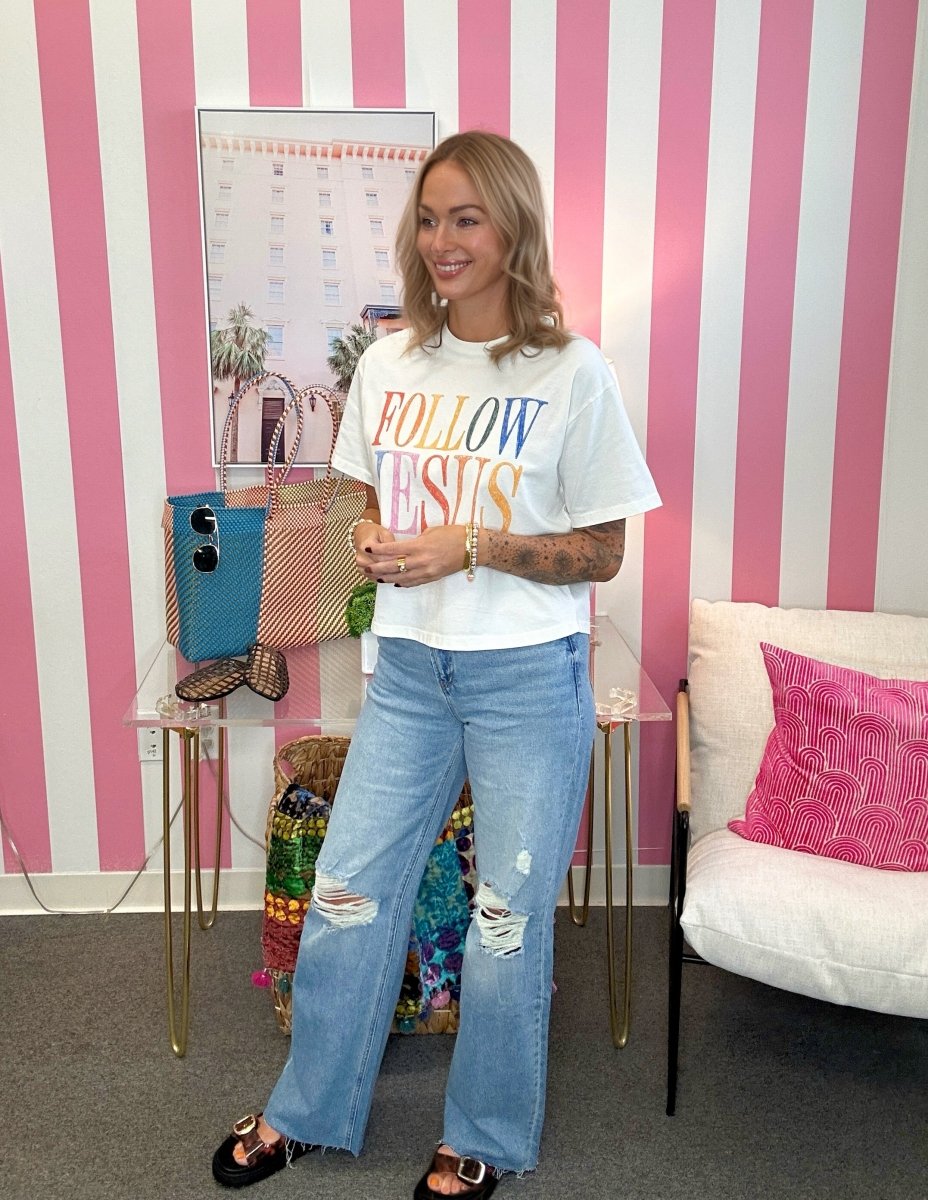A woman stands in a room with pink and white striped walls, wearing a white graphic T-shirt that says 'FOLLOW JESUS' in multicolored letters, light-wash distressed jeans, and black slide sandals.