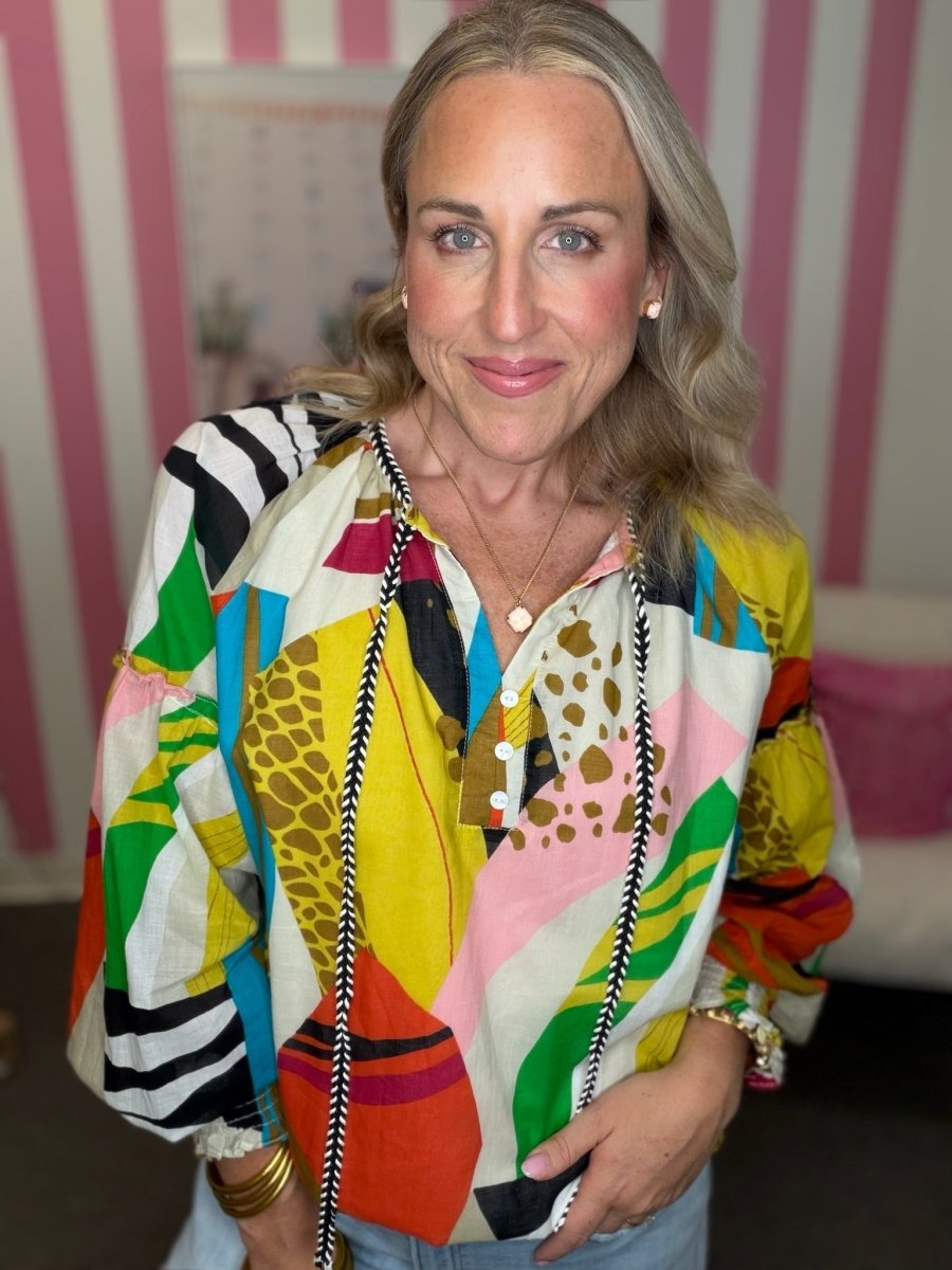 A woman wearing a vibrant, multicolored patchwork print blouse with abstract shapes and animal prints, styled with a long black and white patterned beaded necklace.