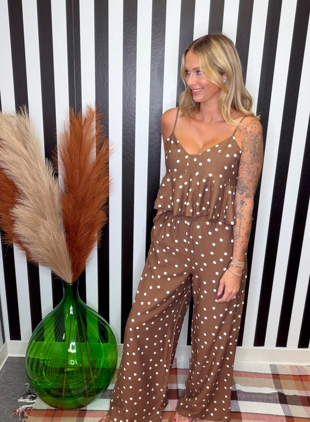 A woman modeling a brown and white polka dot jumpsuit in front of a black and white striped wall, with a green glass vase and pampas grass on the left.