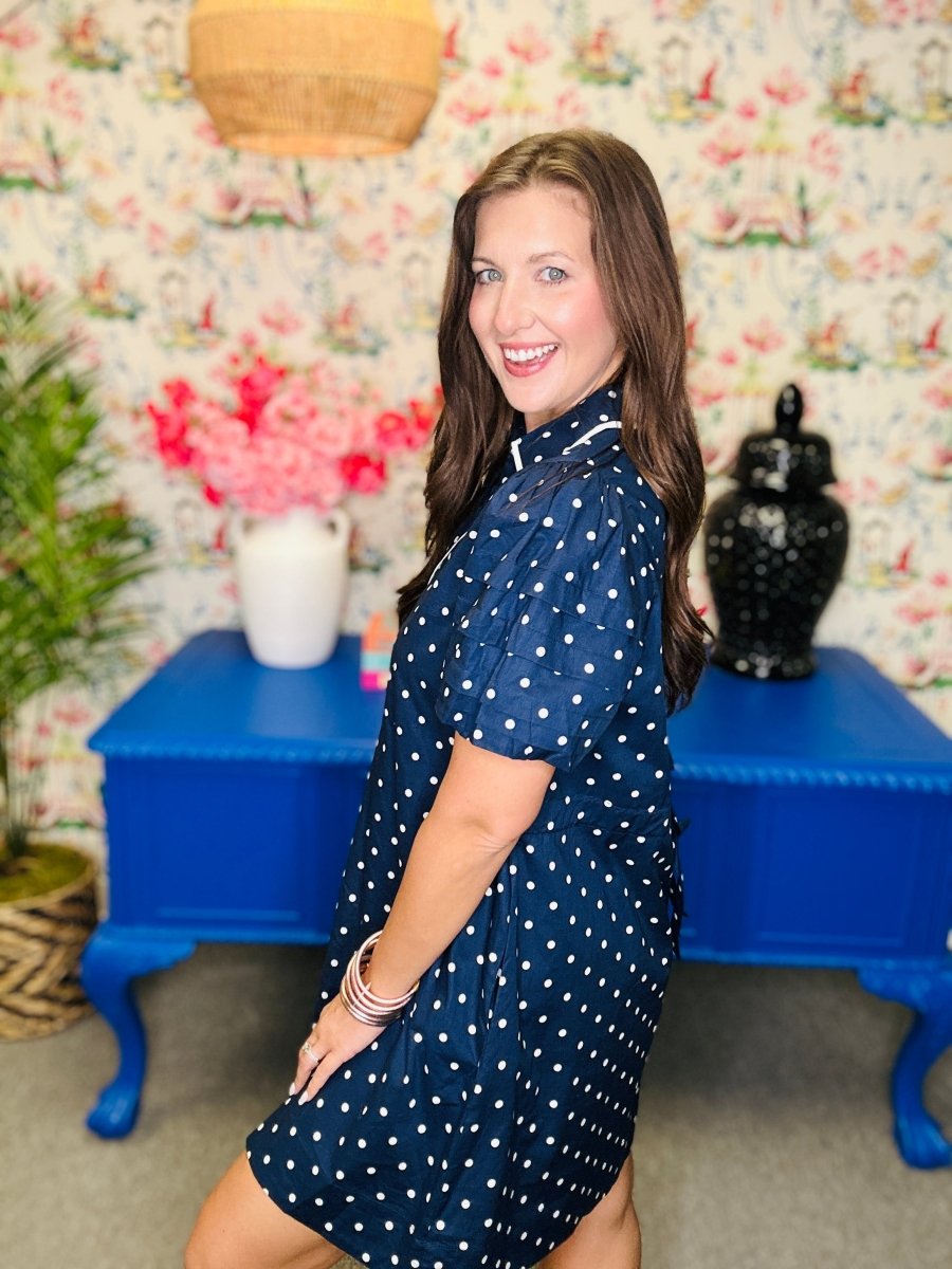 Woman in a blue polka dot dress standing in a room with floral wallpaper and a blue table.