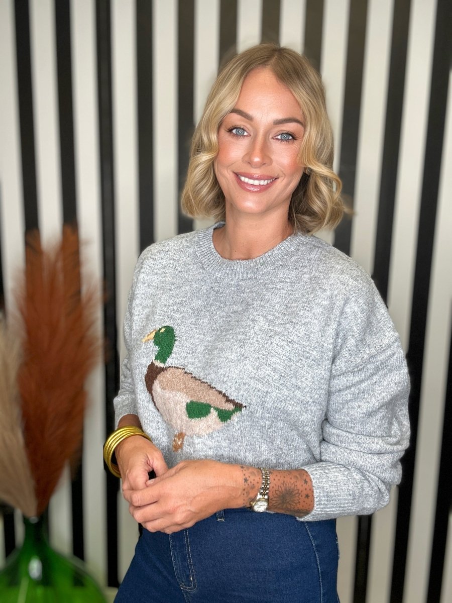 A smiling blonde woman wearing a light gray knit sweater with a large embroidered mallard duck on the front, paired with dark blue jeans. She is standing in front of a black and white vertically striped wall.