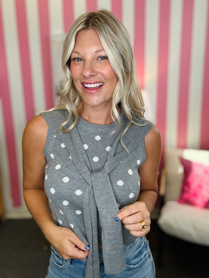A smiling blonde woman wearing a gray sleeveless knit top with white polka dots and a matching knotted scarf, paired with blue jeans, standing in front of a pink and white striped wall.