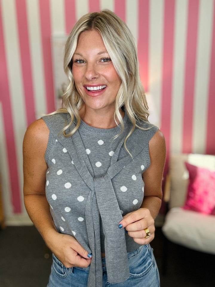 A smiling blonde woman wearing a gray sleeveless knit top with white polka dots and a matching knotted scarf, paired with blue jeans, standing in front of a pink and white striped wall.