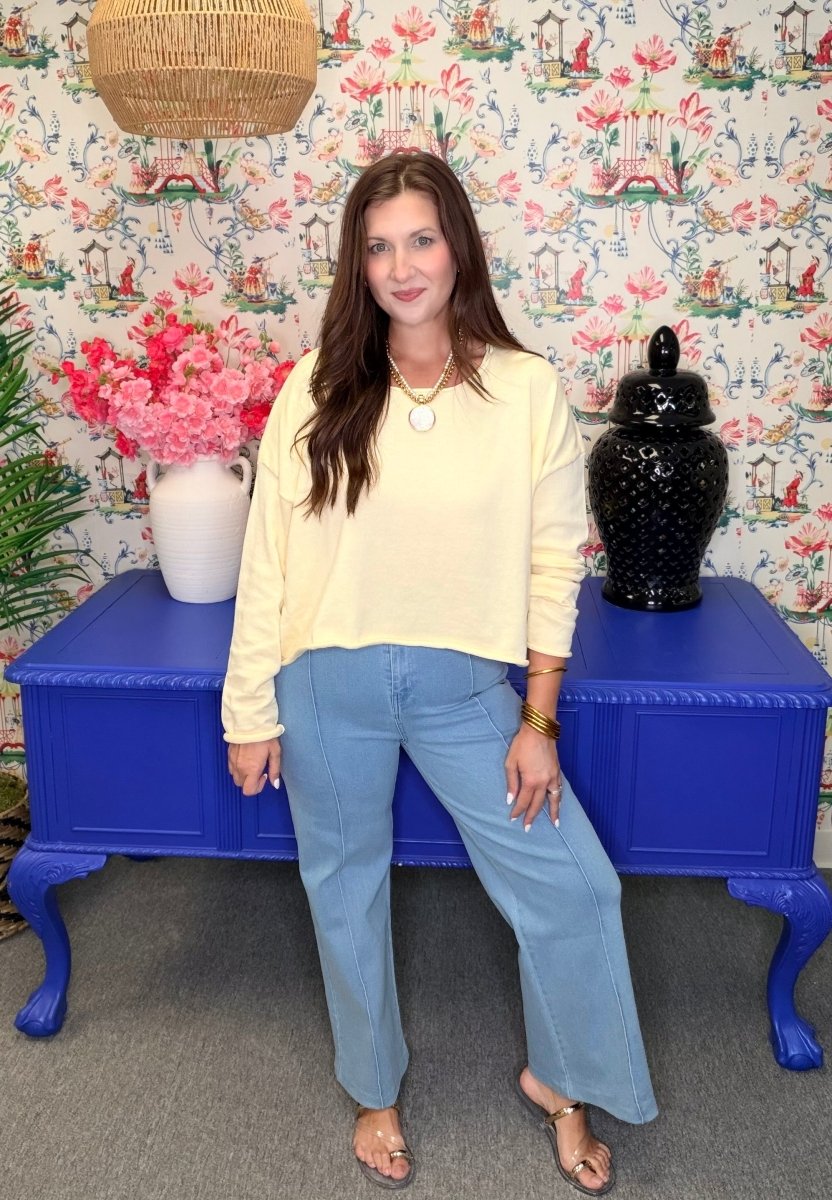 Woman in a yellow top and blue jeans standing in front of a colorful floral-patterned wall with a blue table.