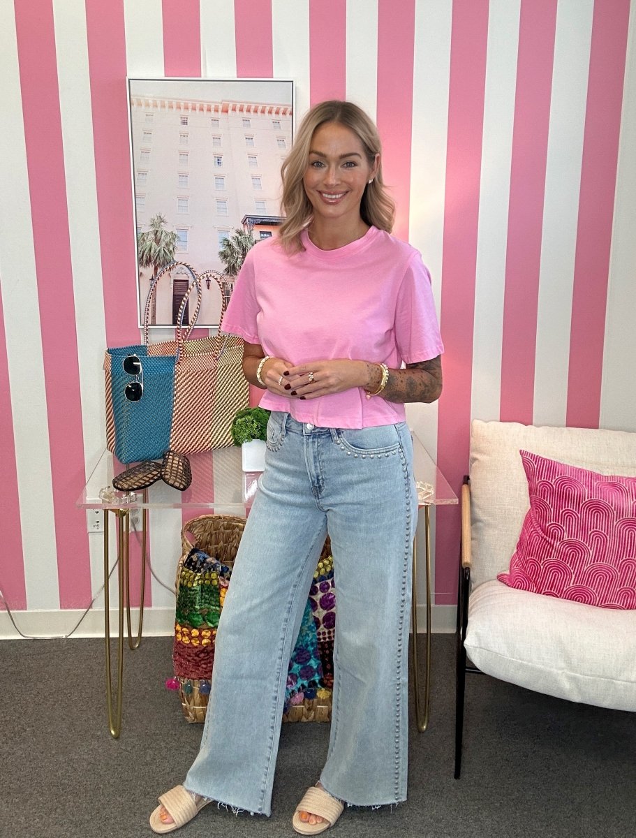A woman stands in a room with pink and white striped wallpaper, wearing a light pink short-sleeve t-shirt and light-wash wide-leg jeans. She is standing next to a glass side table with a blue bag and a white chair with a pink pillow.