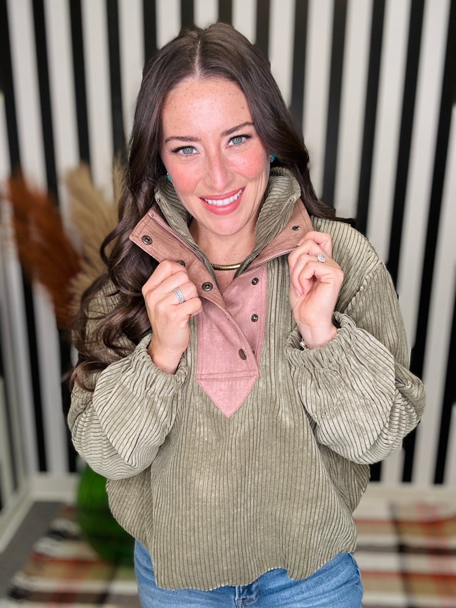 A woman wearing a sage green corduroy pullover with a pink mock neck collar, posing in front of a black and white striped background.