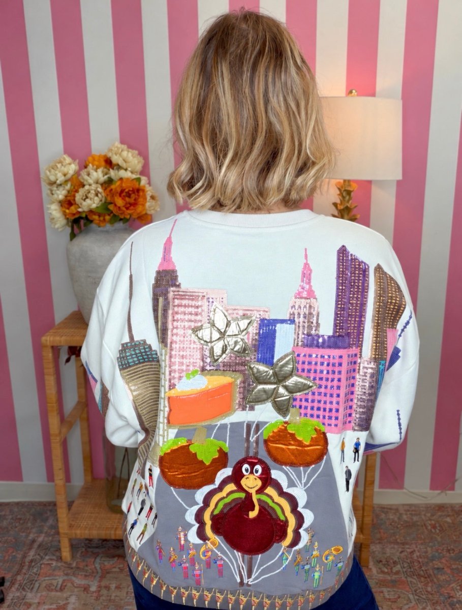A person viewed from behind wearing a white crewneck sweatshirt with a highly detailed, colorful Thanksgiving-themed graphic print featuring a turkey, turkeys, pie, and a city skyline. The background features pink and white vertical stripes, a wooden side table with flowers, and a lamp.