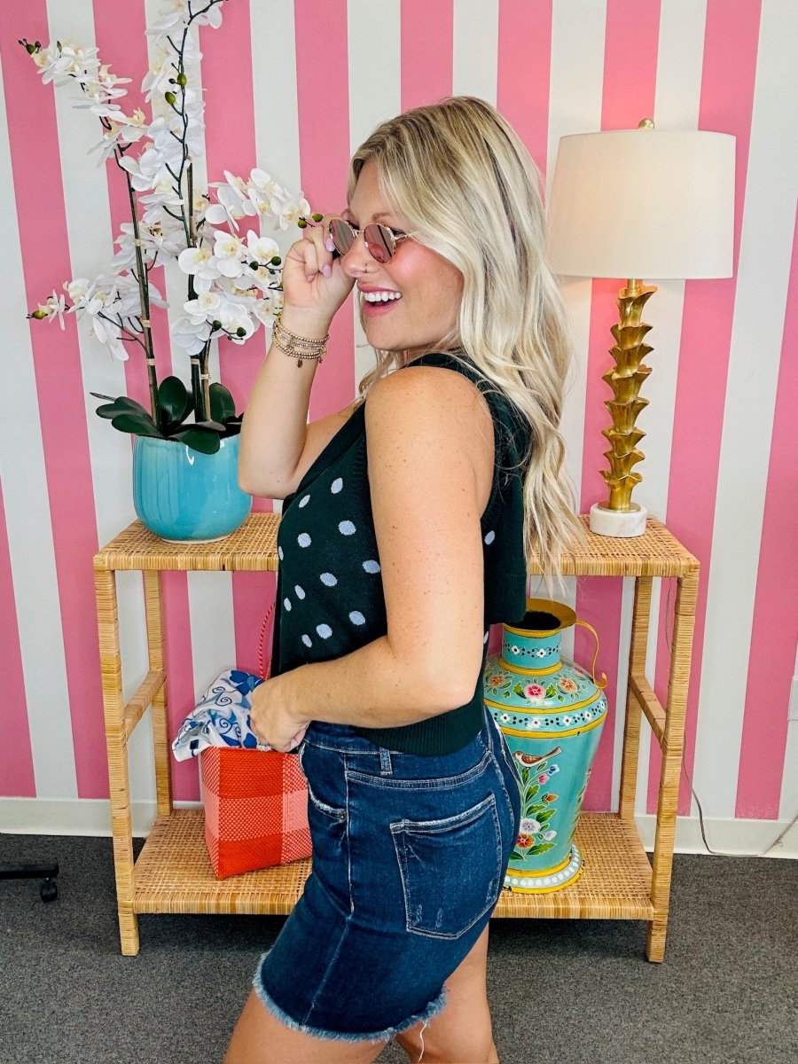 Woman in a polka dot top and denim shorts standing in front of a pink and white striped wall with decorative items.
