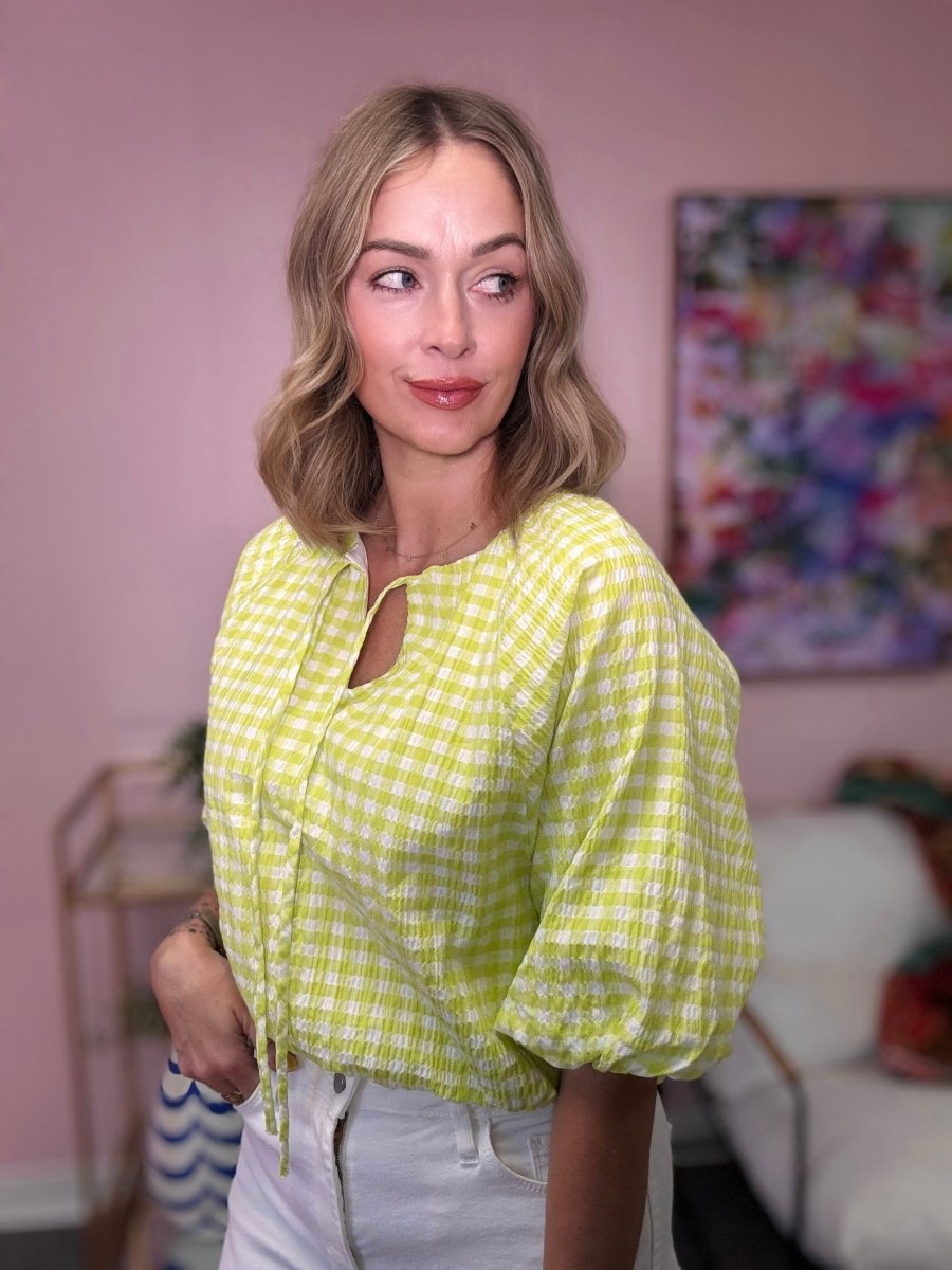 Woman wearing a yellow checkered blouse in a room with a pink wall and colorful artwork.