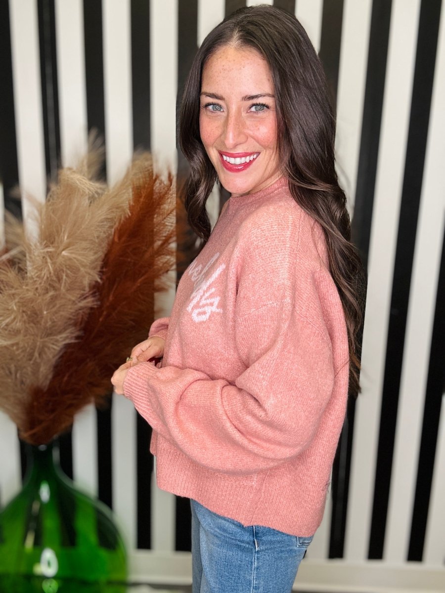 A woman with long dark brown hair smiles while wearing a fuzzy dusty pink crewneck sweater with a white graphic on the chest, paired with blue jeans. She is standing in front of a black and white striped background with a green vase and pampas grass visible on the left.