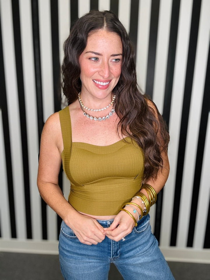 A woman with long wavy brown hair smiling, wearing a ribbed olive green crop top and blue jeans, standing in front of a black and white striped wall.