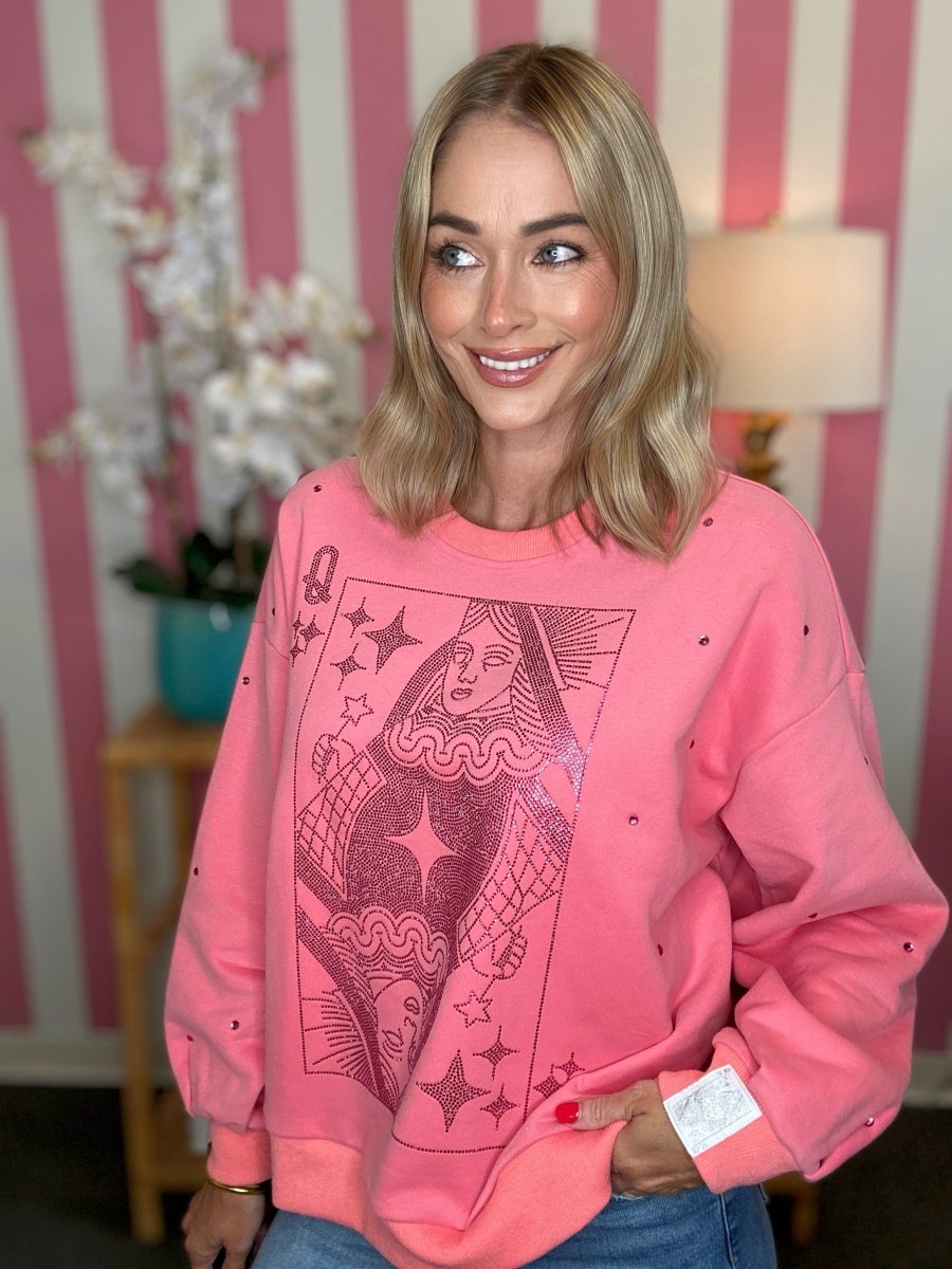 A woman wearing a pink crewneck sweatshirt with a large black playing card graphic and small black dot details, posed in front of a pink and white striped wall.