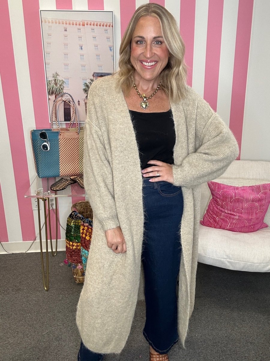A woman stands indoors wearing a long, fuzzy open-front cardigan in a light oatmeal beige color over a black top and dark blue jeans. The background features pink and white vertical stripes, a white sofa with a pink pillow, and a small glass table holding handbags.