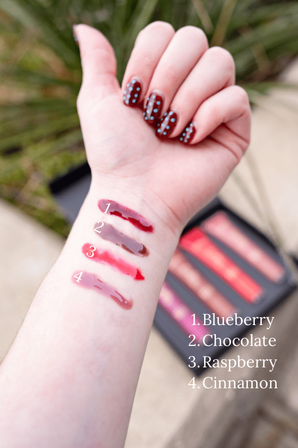 Hand with swatches of lipsticks labeled Blueberry, Chocolate, Raspberry, and Cinnamon against a blurred background.