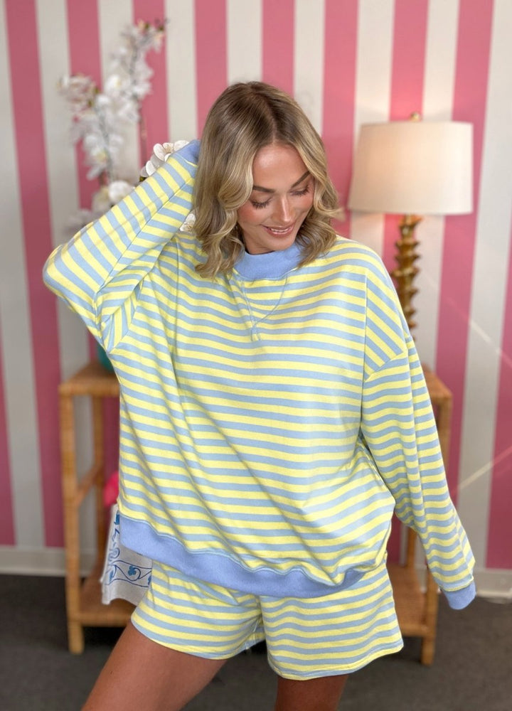 A woman modeling a matching yellow and light blue striped lounge set, featuring a long-sleeve sweatshirt and shorts, posed in front of a pink and white striped wall.