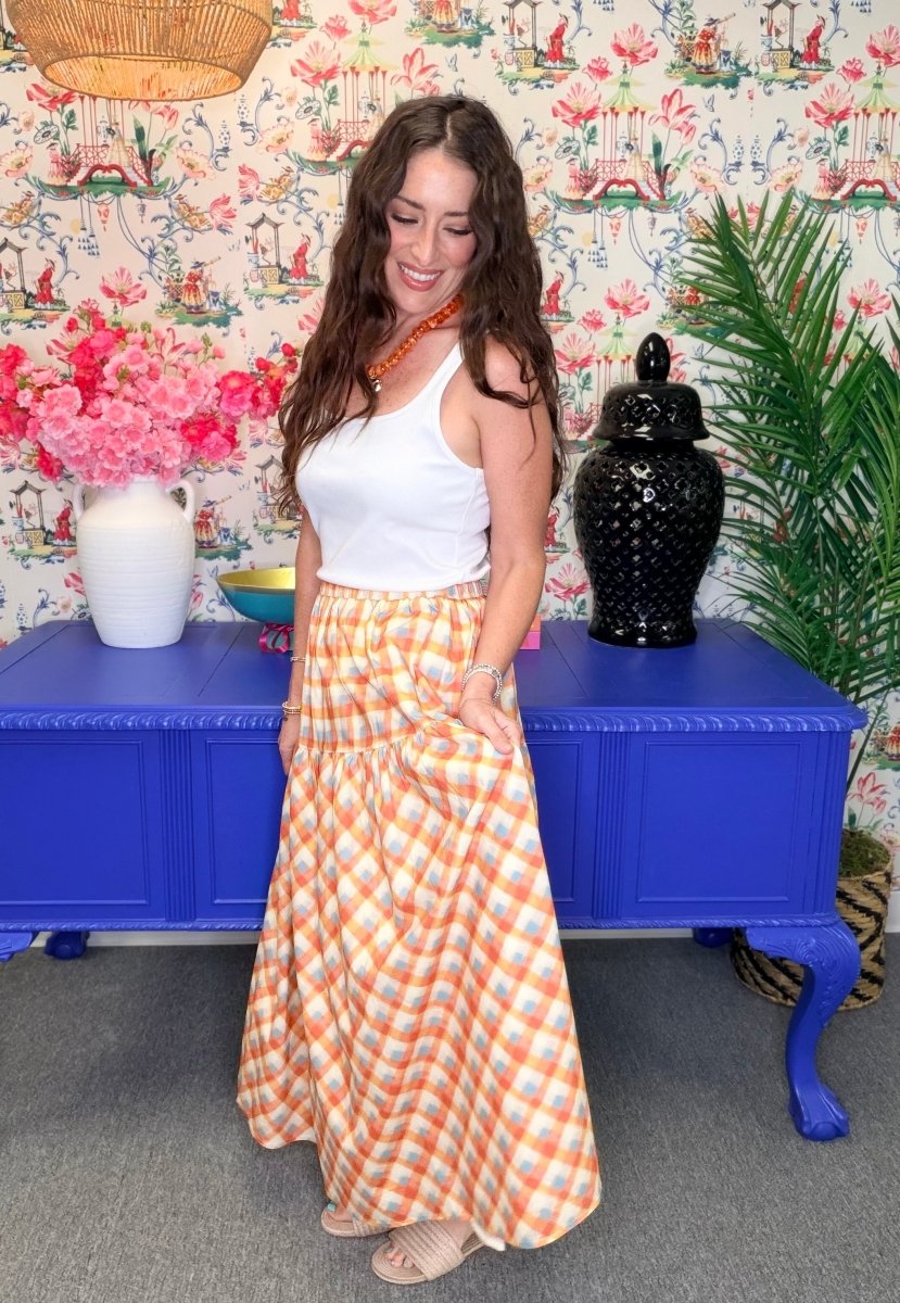 A woman modeling a white ribbed tank top and a long orange and white gingham maxi skirt, standing in front of a blue cabinet and a floral wallpaper background.