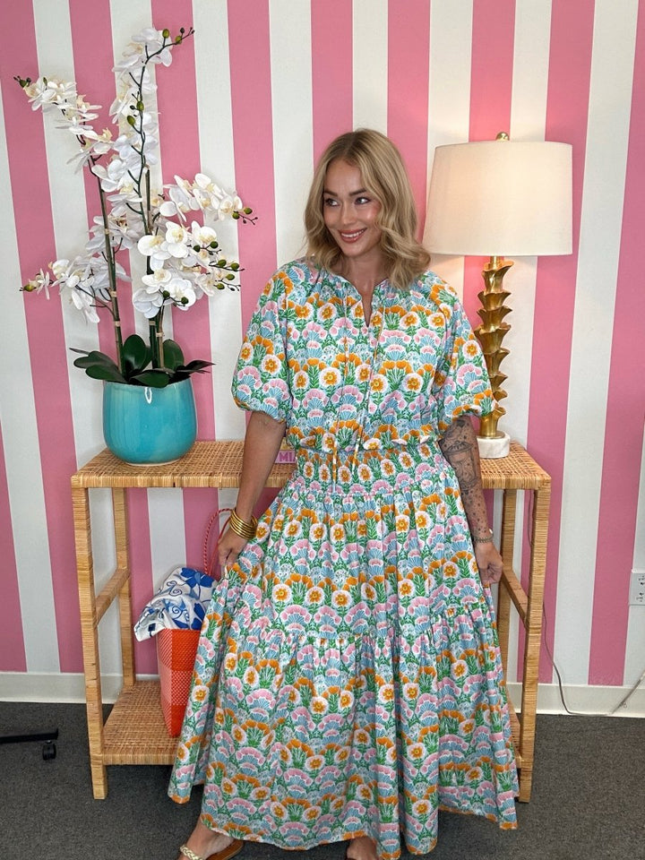 Woman in a floral dress standing in front of a pink and white striped wall with a lamp and plant.