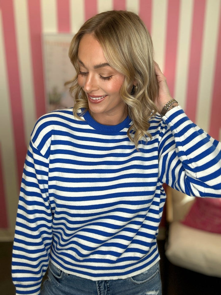 A woman wearing a blue and white horizontally striped long-sleeve knit top, paired with blue jeans, standing in front of a pink and white striped wall.