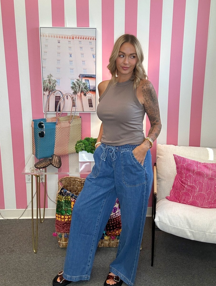 A woman stands in a room with pink and white striped walls, wearing a sleeveless taupe top and wide-leg blue jeans. She is posing next to a white sofa with a pink pillow and a small side table holding colorful bags and accessories.