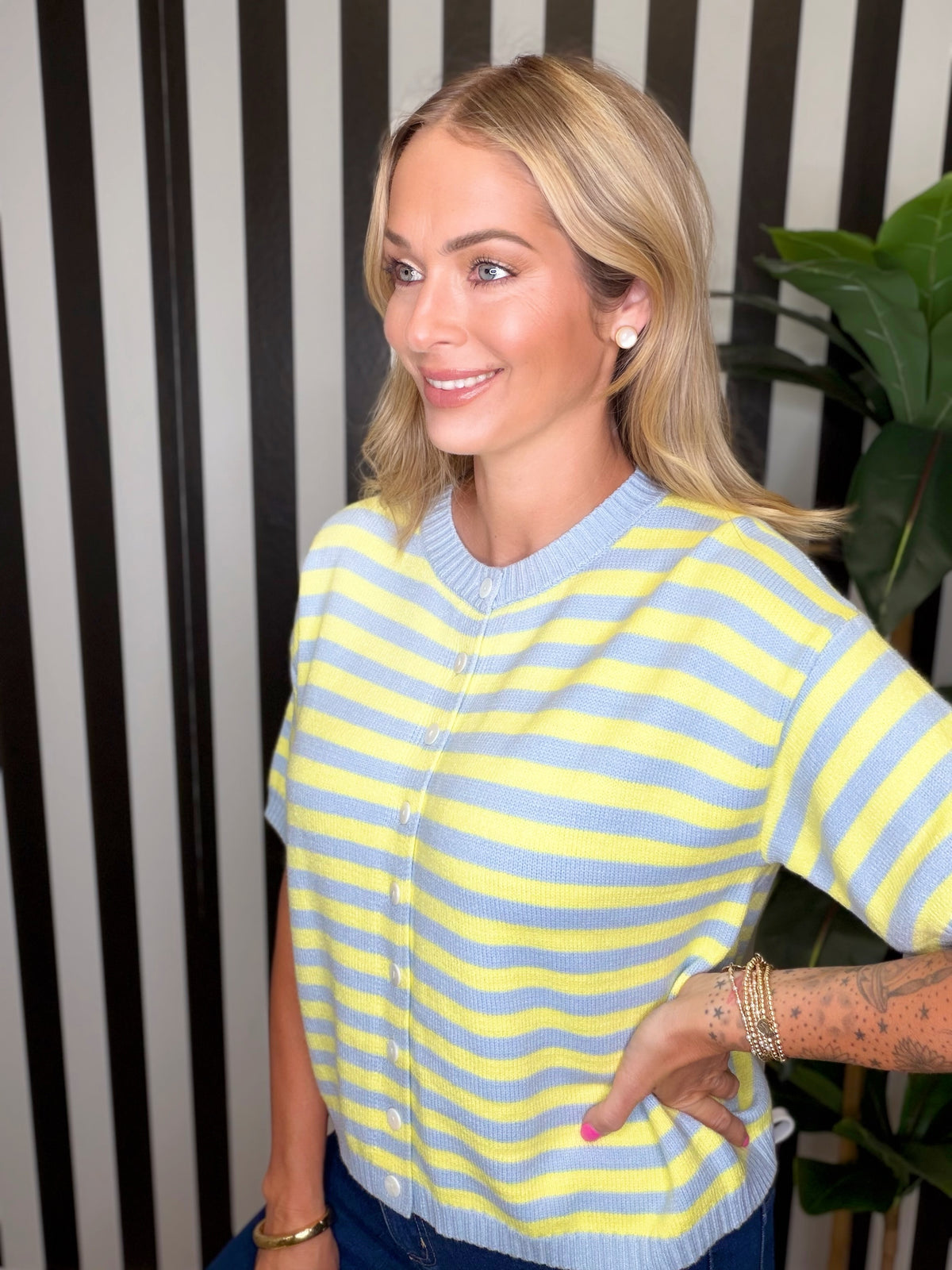 A woman wearing a yellow and light blue striped short-sleeve knit cardigan sweater, standing in front of a black and white striped wall with a green plant in the background.