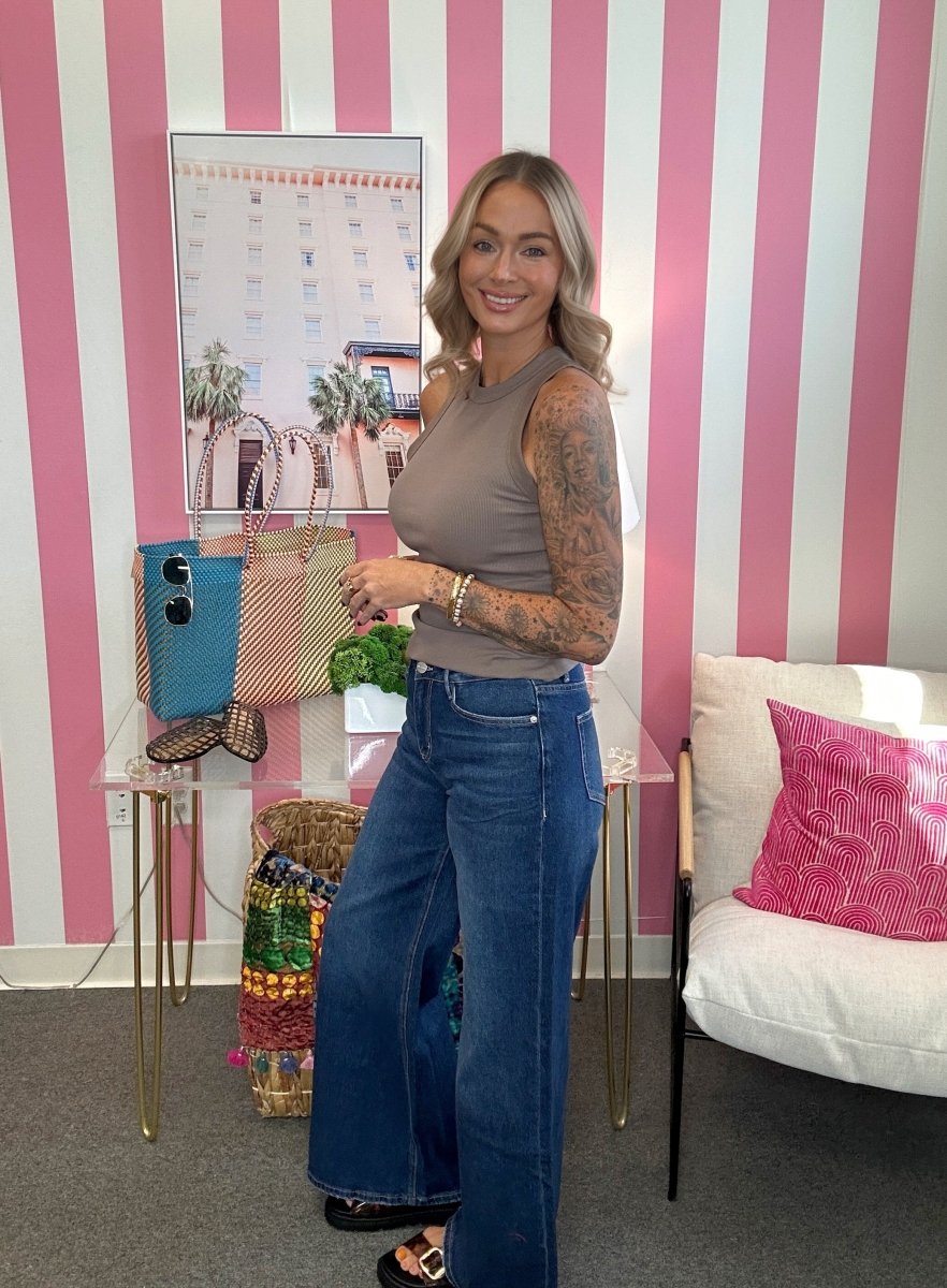 A woman with blonde hair and tattoos stands in a room with pink and white striped walls, wearing a taupe sleeveless top and wide-leg blue jeans. She is posing next to a glass table holding a blue and pink woven tote bag.