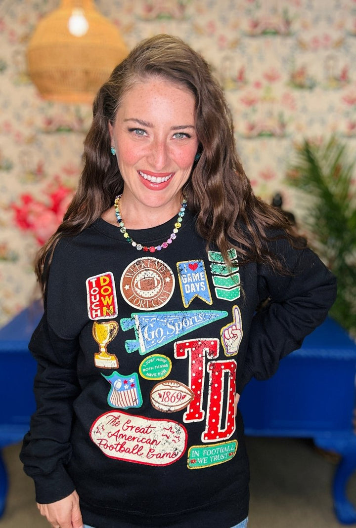 A smiling woman with long brown hair wears a black long-sleeve sweatshirt covered in colorful football-themed patches and graphics, including 'TD', 'Peanut Butter', 'Cheddar', and 'The Great American Football Crawl'. She is standing indoors against a floral wallpaper background with a blue table visible behind her.