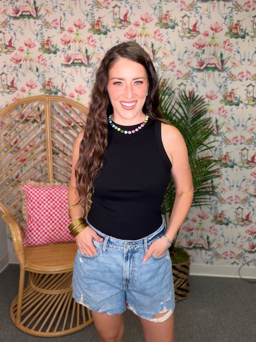 Woman in black sleeveless top and denim shorts standing in a room with floral wallpaper.