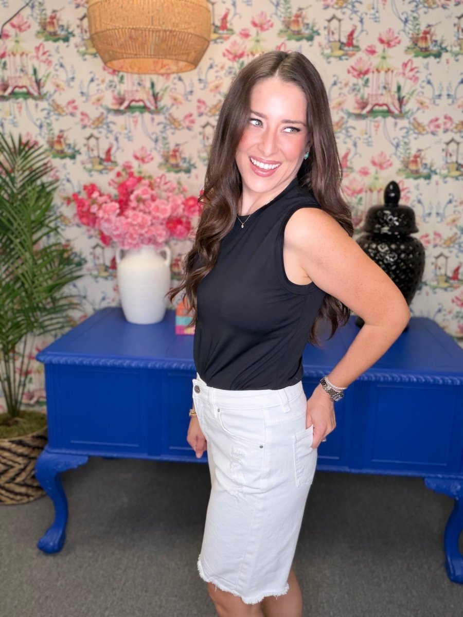 Woman in black sleeveless top and white shorts standing in a room with floral wallpaper and a blue table.