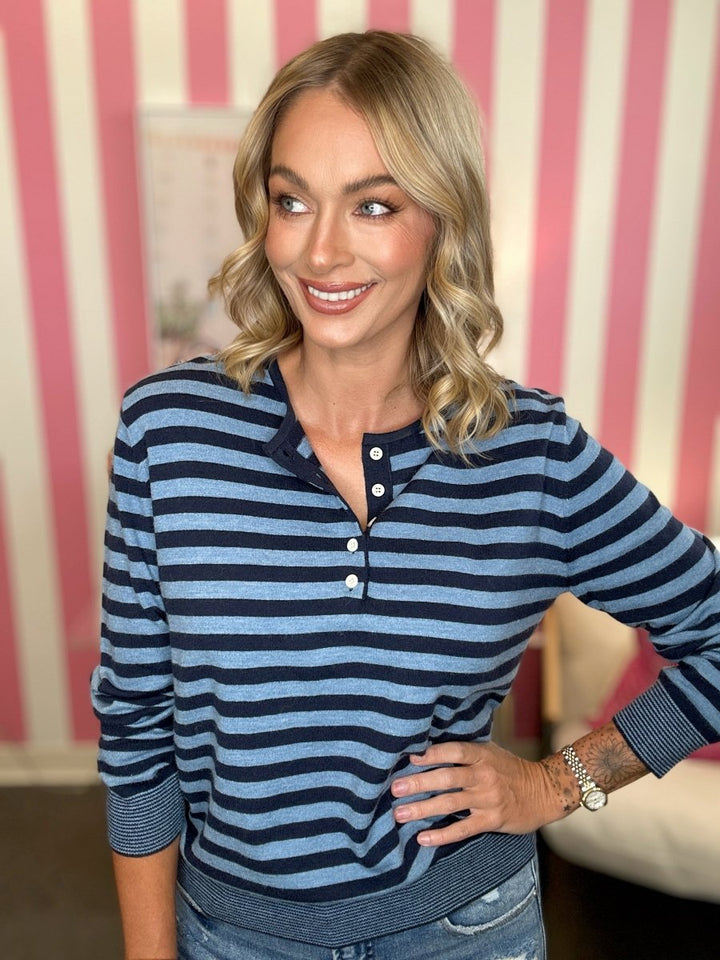 A woman wearing a navy and light blue striped long-sleeve henley shirt with three buttons, paired with blue jeans, standing in front of a pink and white striped wall.