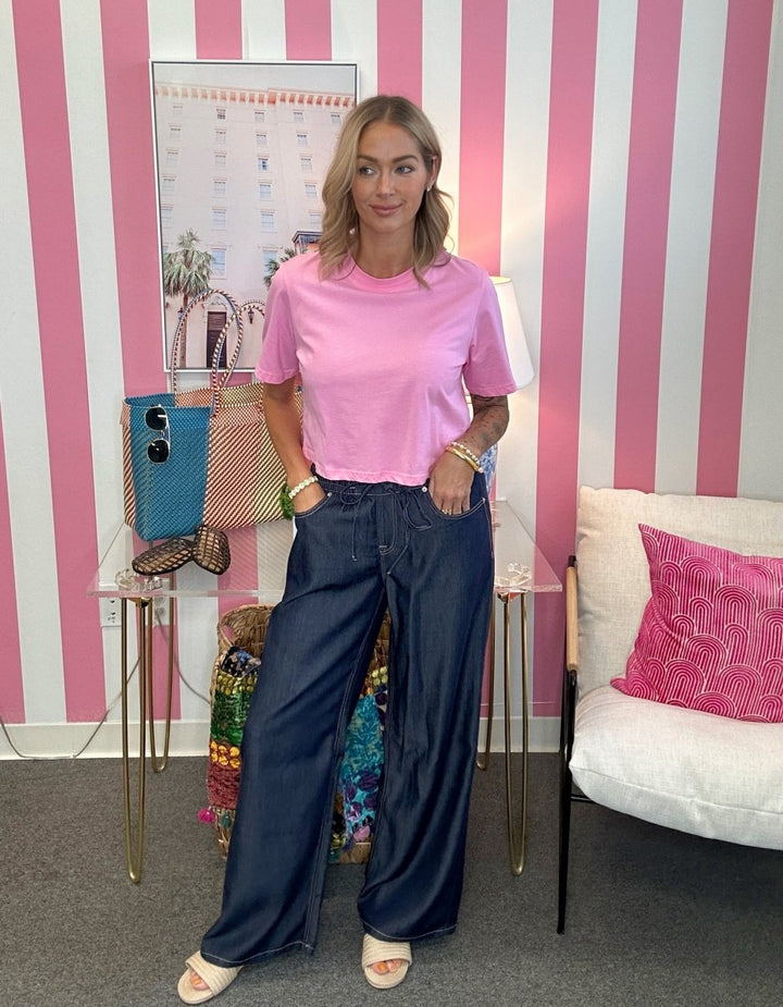 A woman stands in a room with pink and white vertical stripes, wearing a pink short-sleeve t-shirt and dark blue wide-leg jeans. She is standing next to a white armchair with a pink pillow and a small table holding a blue handbag and a patterned bag.
