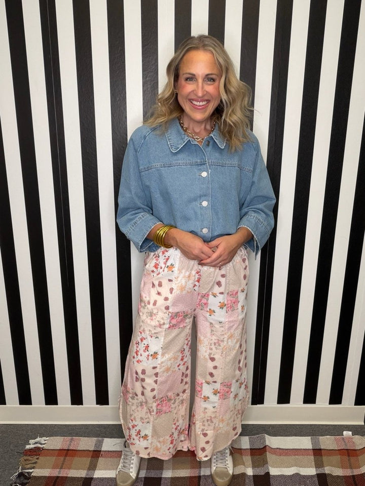A woman stands in front of a black and white striped wall, wearing a light blue denim shirt and wide-leg cream pants with a pink and orange floral pattern. She is wearing gold bracelets and white sneakers.