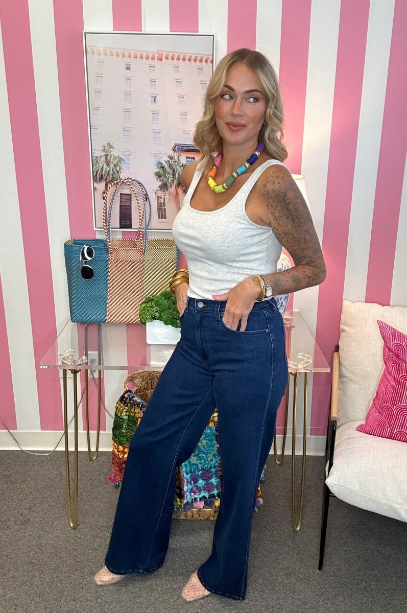 A woman stands in a room with pink and white vertical stripes, wearing a white ribbed tank top, dark blue wide-leg jeans, and a colorful beaded necklace.