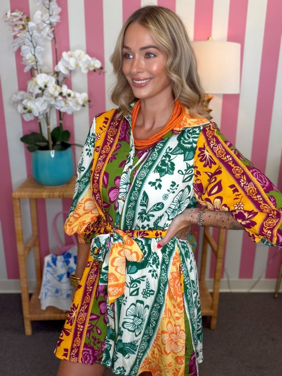 A woman modeling a colorful patchwork-print kaftan dress with yellow, green, orange, and white panels, standing in front of a pink and white striped wall.