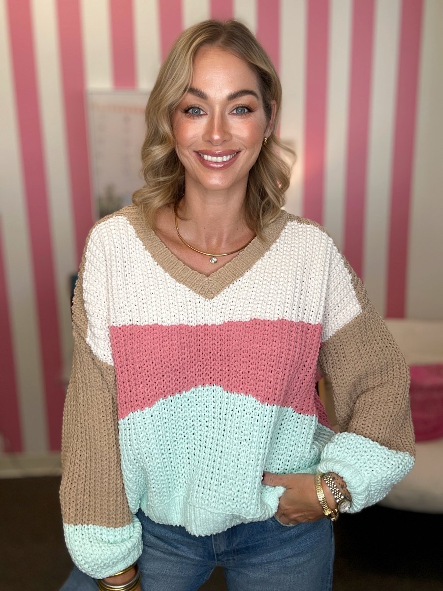 A woman wearing a chunky knit color-block sweater with beige, white, pink, and mint green stripes, paired with blue jeans, standing in front of a pink and white striped wall.
