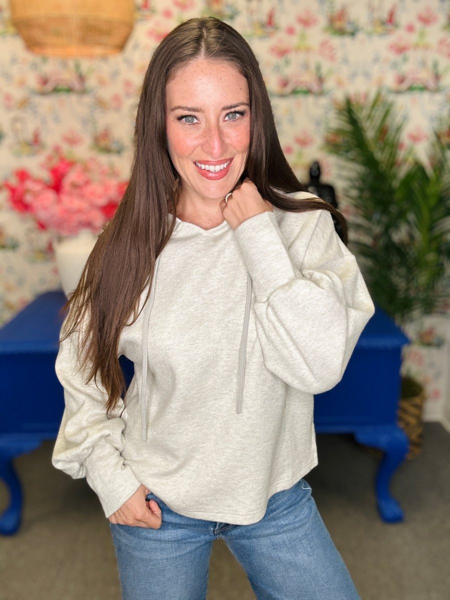 A woman with long brown hair wearing a light beige heathered pullover hoodie and blue jeans, posing in a decorated room with floral wallpaper, a blue table, and a green plant.
