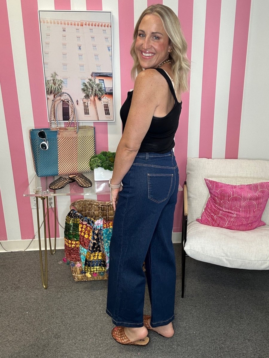 A woman modeling a black tank top and dark blue wide-leg jeans, standing in a room with pink and white vertical stripes on the wall, a white chair with a pink pillow, and a small glass table holding colorful handbags.