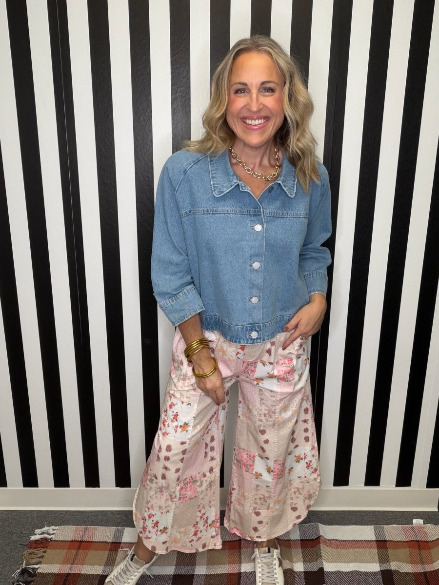A woman stands in front of a black and white striped wall wearing a light blue denim button-up shirt and light pink wide-leg pants with a floral print. She is wearing gold jewelry and white sneakers.
