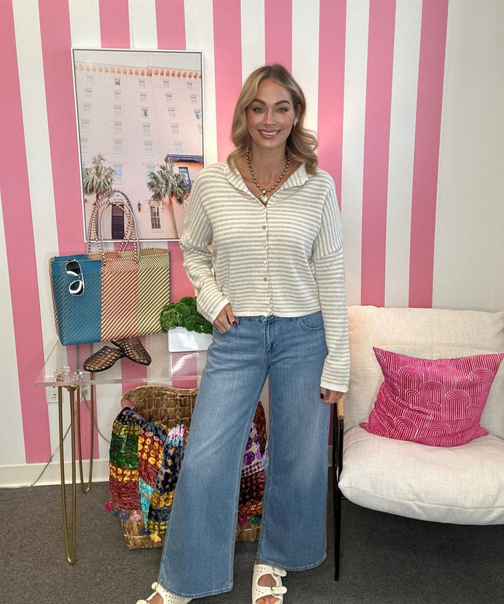 A woman stands in a room with pink and white vertical stripes, wearing a cream and white striped long-sleeve button-up shirt and wide-leg blue jeans. She is accessorized with layered gold necklaces and white heeled sandals.