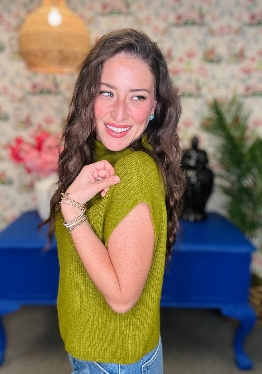 A woman with long wavy brown hair smiling and posing in a ribbed olive green sleeveless top, set against a background of floral wallpaper, a blue table, and a hanging lamp.