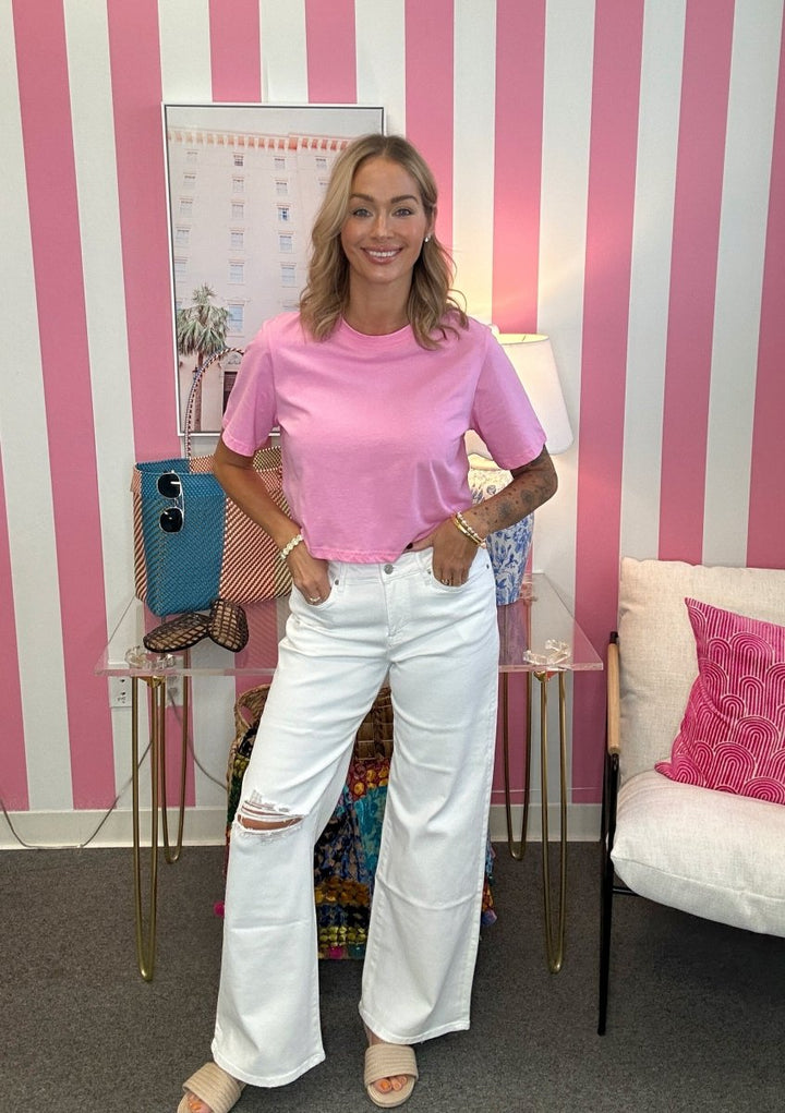 A woman stands in a room with pink and white striped wallpaper, wearing a light pink short-sleeve crop top and white wide-leg distressed jeans.
