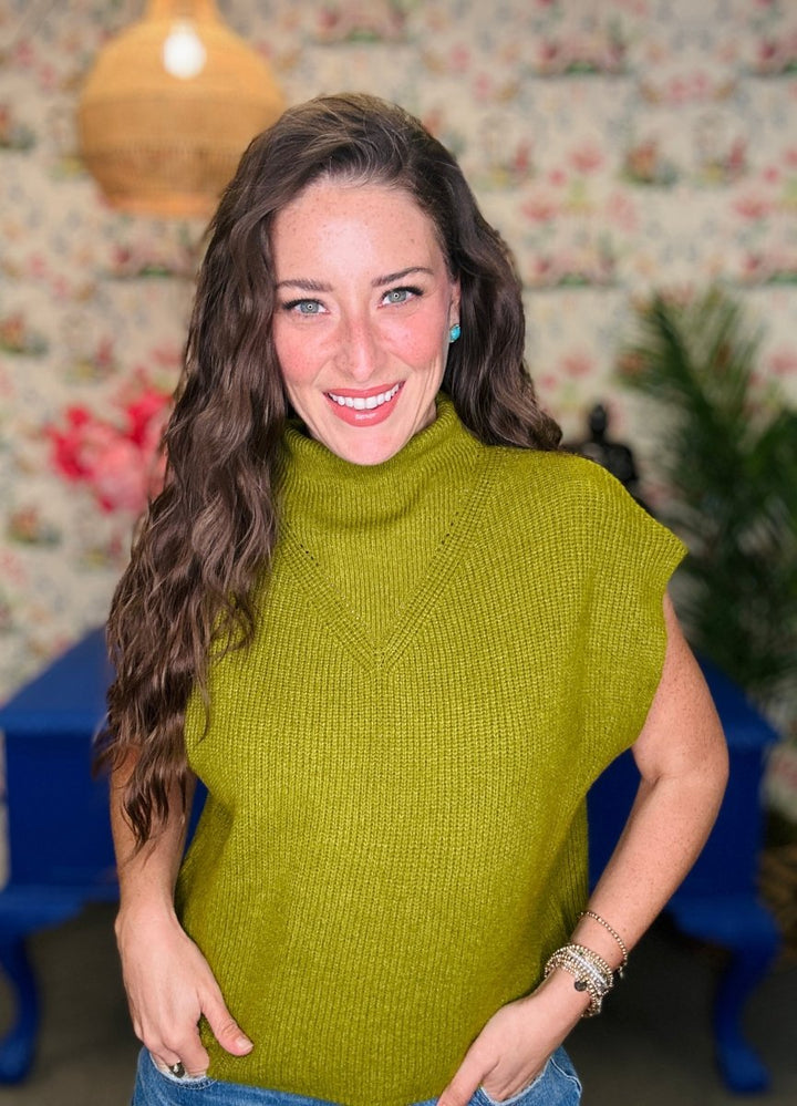 A smiling woman with long wavy brown hair wearing a ribbed olive green mock neck top with short sleeves, paired with blue jeans, standing in a room with floral wallpaper and a blue chair in the background.