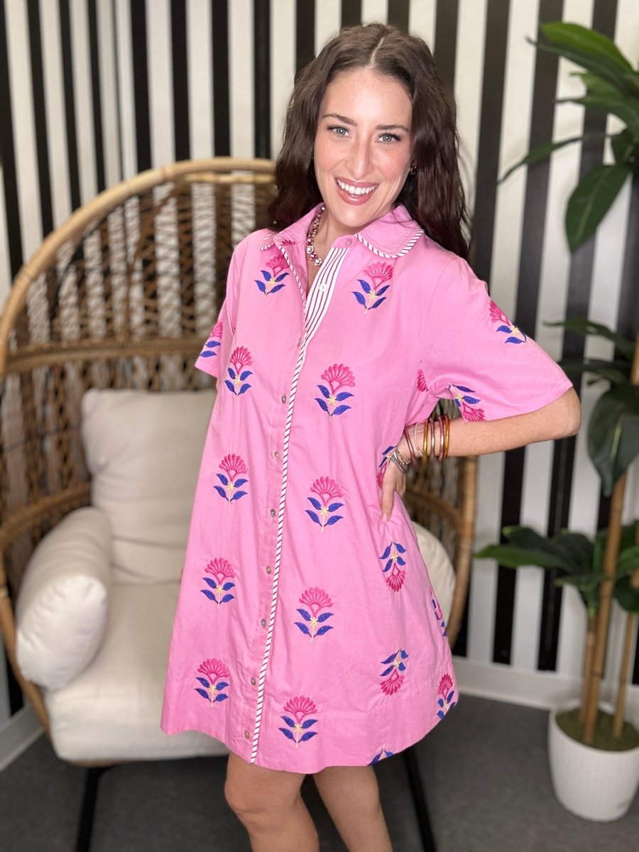 Woman wearing a pink dress with floral patterns in a room with a striped wall and wicker chair.