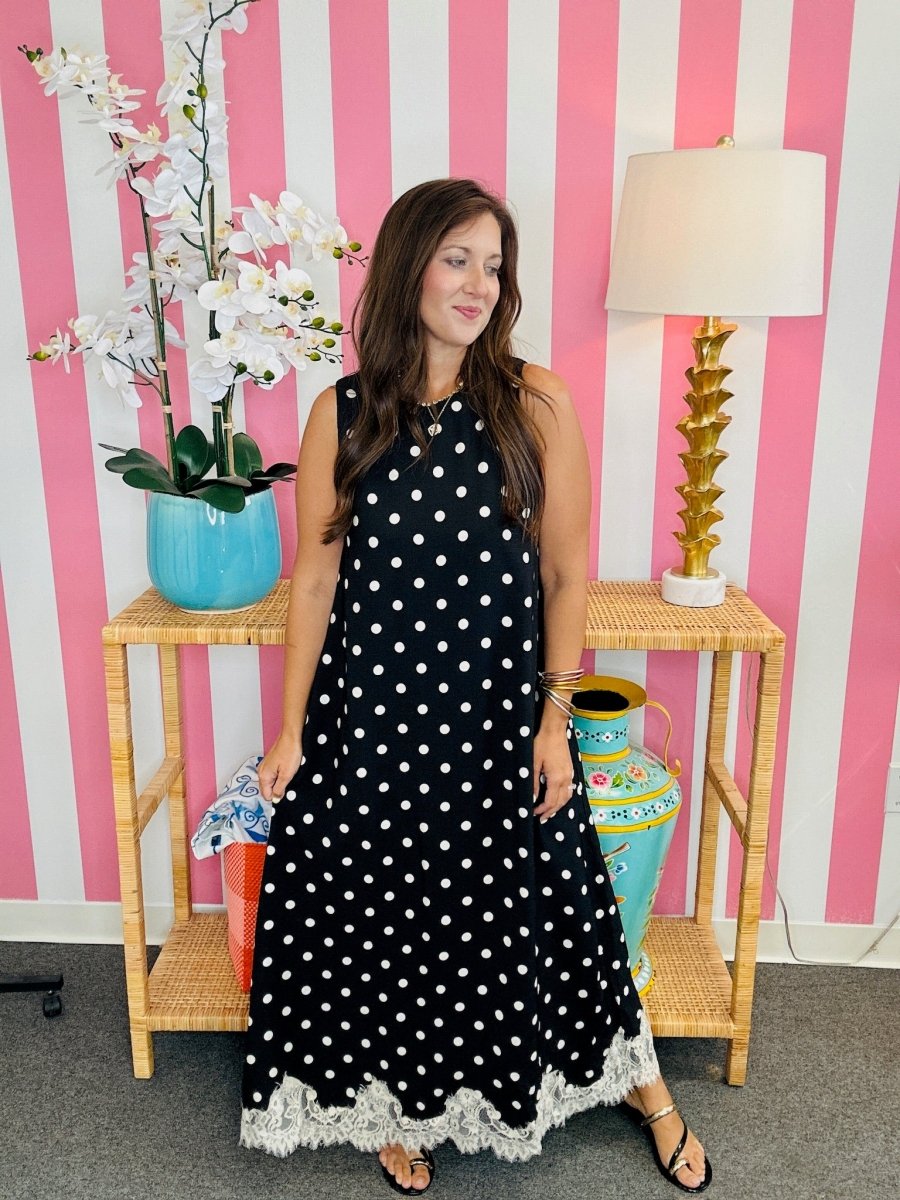 A woman wearing a sleeveless black maxi dress with white polka dots and a white lace hem, standing in front of a pink and white striped wall. She is positioned between two light wood side tables with decorative items, including a blue vase with white flowers, a gold lamp, and a colorful ceramic pot.