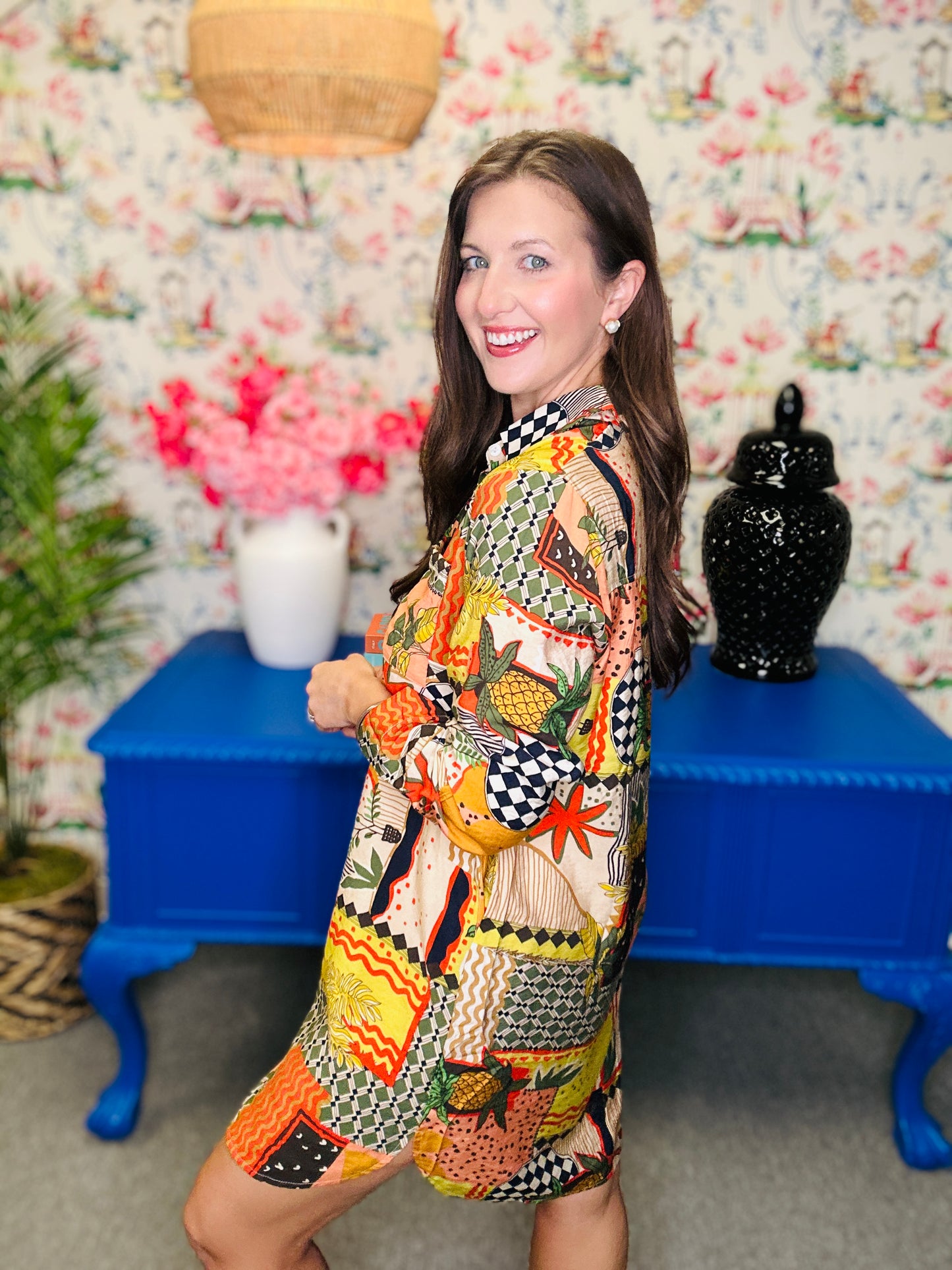 Woman in a colorful patterned dress standing in front of a floral wall with a blue table and decorative items.