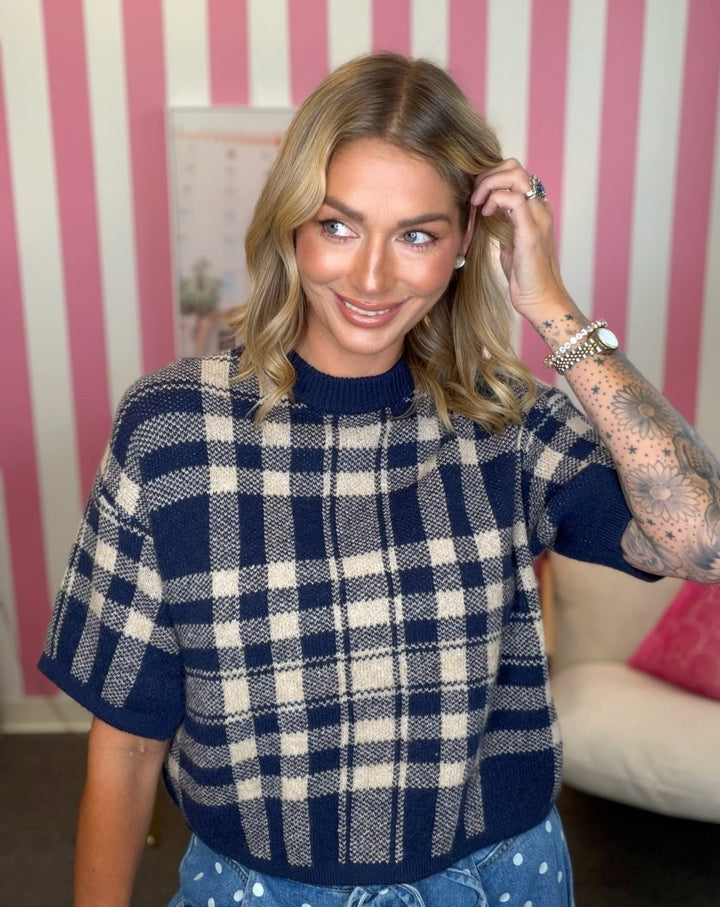 A woman wearing a navy and cream plaid short-sleeve knit sweater, standing in front of a pink and white striped wall.