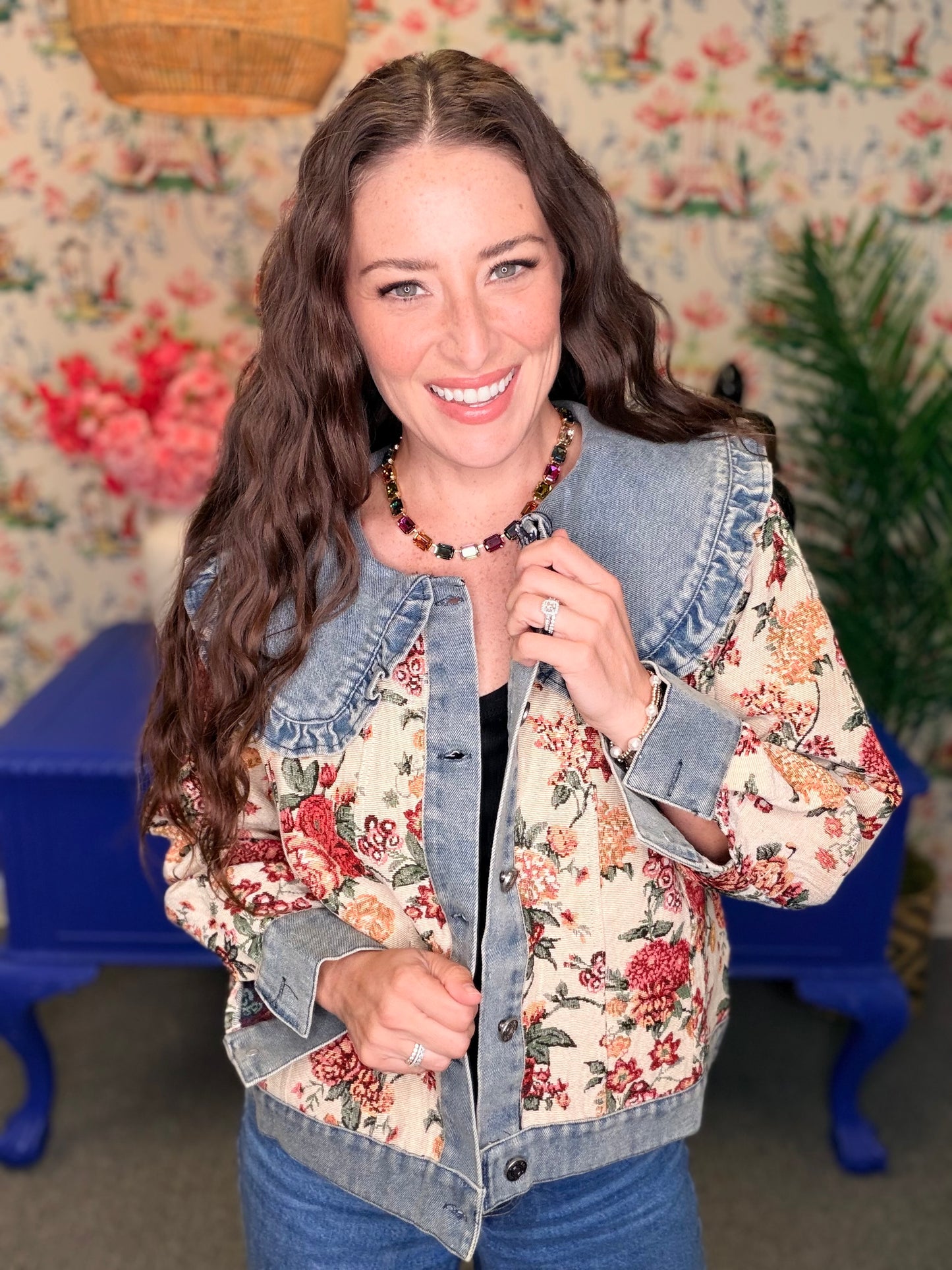 A woman smiling and wearing a denim jacket with a red and orange floral print on the front panels, set against a vintage floral wallpaper background with a blue table.