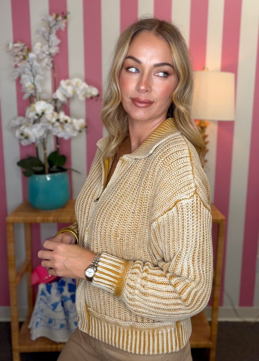 A woman wearing a chunky knit zip-up cardigan sweater in a beige and mustard yellow colorway, standing in front of a pink and white striped wall.