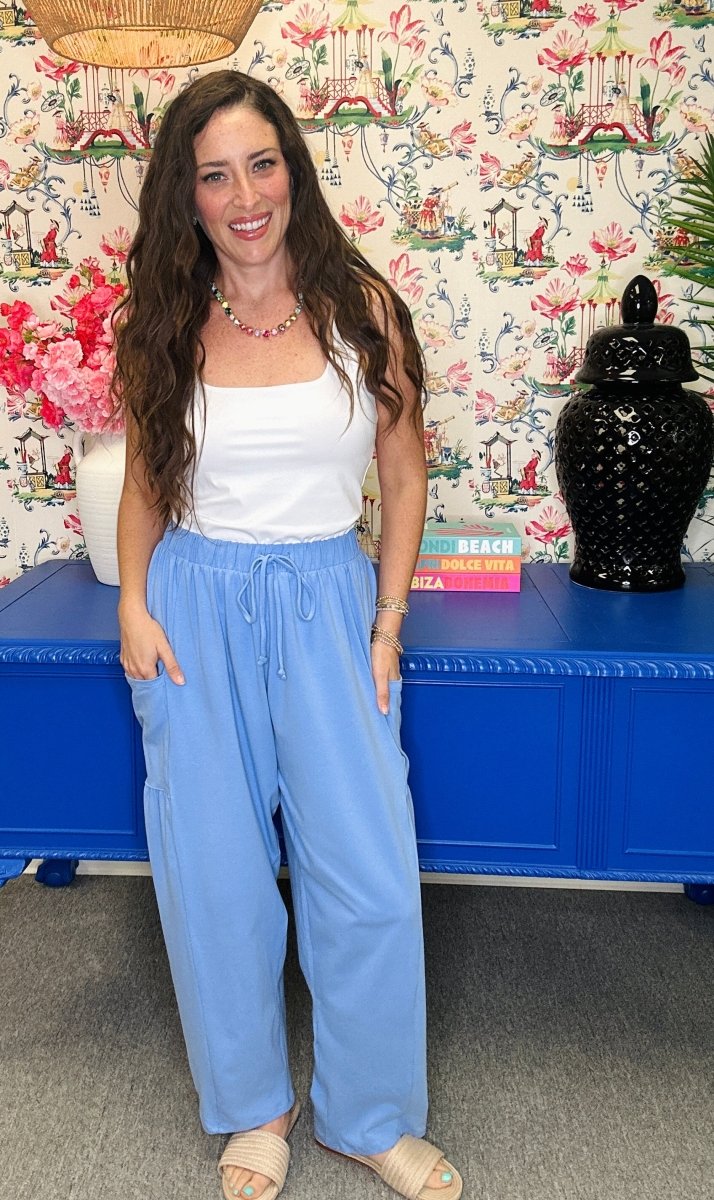 A woman stands indoors wearing a white sleeveless top and light blue wide-leg drawstring pants, with a floral wallpaper background and a blue cabinet behind her.