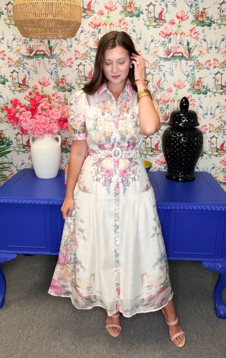 Woman in a floral dress standing in front of a colorful wall with a blue table and decorative items.