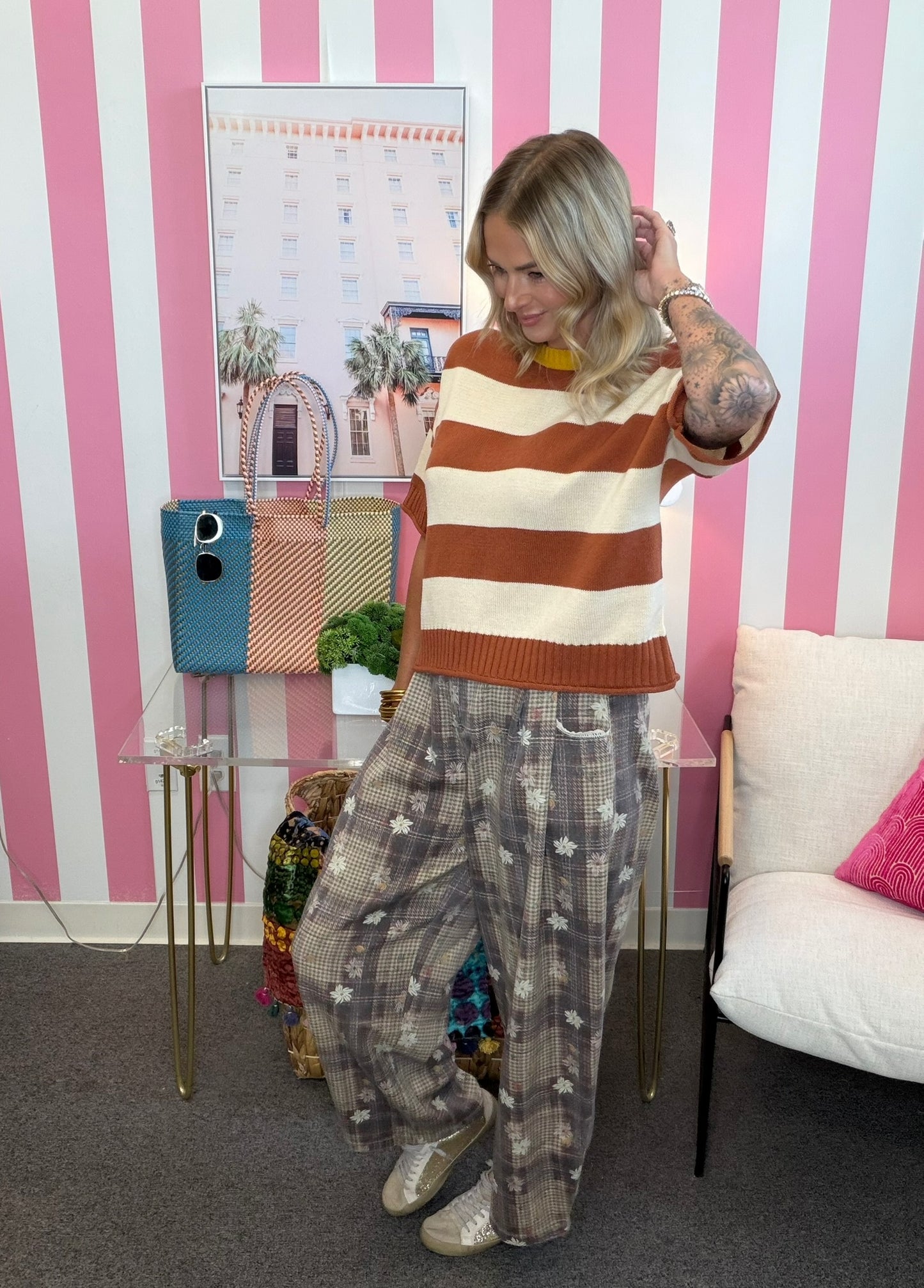 A woman modeling a brown and cream striped knit sweater and grey patterned wide-leg trousers, standing in front of a pink and white striped wall.